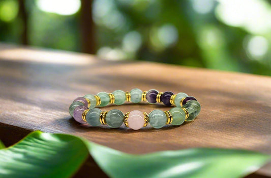 A bracelet with Rainbow Fluorite and Rose Quartz beads, gold-colored metal accents, displayed against a leafy background.